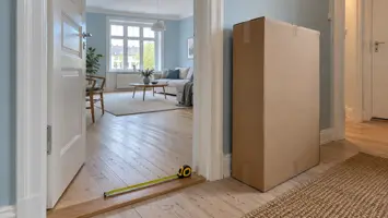 Illustration: Døråbning, målebånd og papkasse i en entré